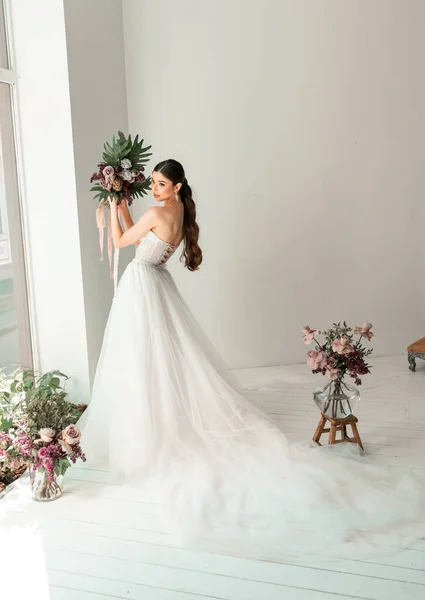 Vacker brud poserar i bröllopsklänning i en vit fotostudio. Bröllopsbukett med blommor — Stockfoto