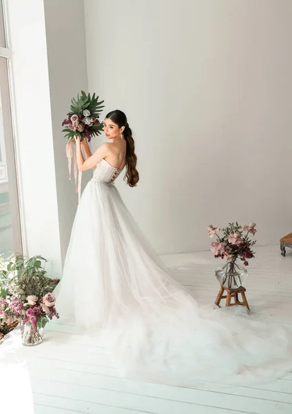 Vacker brud poserar i bröllopsklänning i en vit fotostudio. Bröllopsbukett med blommor Stockfoto