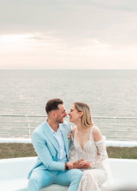 Güzel bir düğün çifti, damat ve gelin, deniz kıyısında dinleniyorlar, kucaklaşıyorlar, el ele tutuşuyorlar, eğleniyorlar. Düğün istirahatı, balayı konsepti. Günaydın, gündoğumu