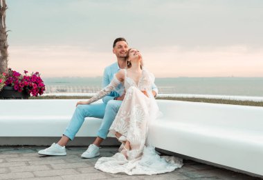 Güzel bir düğün çifti, damat ve gelin, deniz kıyısında dinleniyorlar, kucaklaşıyorlar, el ele tutuşuyorlar, eğleniyorlar. Düğün istirahatı, balayı konsepti. Günaydın, gündoğumu