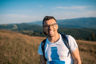 Kapalı gözlü yakışıklı sakallı adam kameraya gülüyor ve dilini gösteriyor. İnsan Gezgin, bir dağ manzarasının kamera arkasında duygularını gösterir. Yolculuk Konsepti