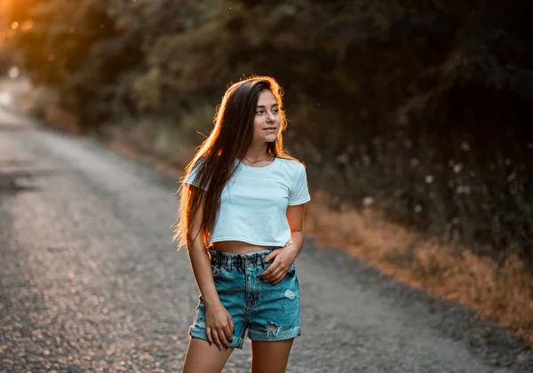 Açık havada poz veren esmer genç kız. - Yakın çekim. Beyaz tişörtlü, mavi şortlu, günbatımı arka planında poz veren modaya uygun bir kadın. Çapraz kollar