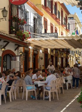 İnsanlar Ronda 'da (Endülüs, İspanya) bir barın terasında tapas yiyip içiyorlar. Sokak bar teraslarıyla dolu. Sokakta bir sürü insan oturuyor, yemek yiyor ya da tapas yiyor..