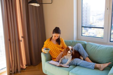 Genç bir kadın kanepesinde Cavalier King Charles Spaniel ile oturuyor ve kitap okuyor..