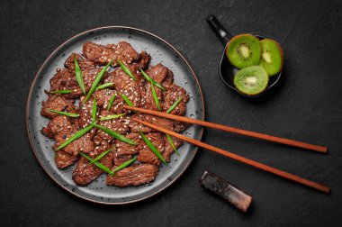 Beef Bulgogi on black plate on dark slate table top. Korean cuisine meat dish with sauce and kiwi. Traditional asian food. Top view