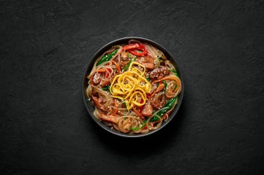 Japchae in black bowl on dark slate table top. Korean cuisine glass chapchae noodles dish with vegetables and meat. Asian traditional food. Authentic meal. Top view