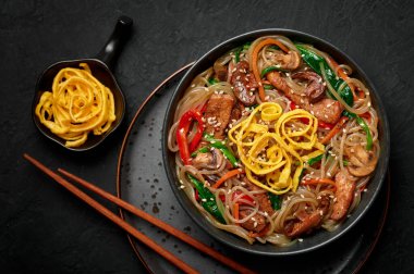 Japchae in black bowl on dark slate table top. Korean cuisine glass chapchae noodles dish with vegetables and meat. Asian traditional food. Authentic meal. Top view