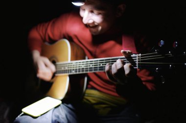 Young tourist man playing guitar, singing and using phone app with chords and song lyrics. Guitarist traveler in the night singing a song.