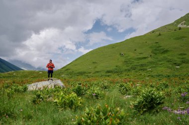 Young tourist man with a camera stands on the big stone among the green hills in the mountains.