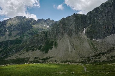 High mountain range landscape with rocky hills, river, green valley and white clouds.