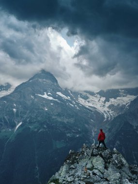 Alone hiker man in red jacket against the gloomy mountain top landscape with thunder cloudy sky, rocky ranges and peaks with glaciers and snow fields. Domestic travel and trekking. Local tourism.