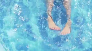 Havuzda ayaklarıyla oynayan çocuk yaz tatilinde havuzda yüzerken mavi su sıçratmayı öğreniyor.