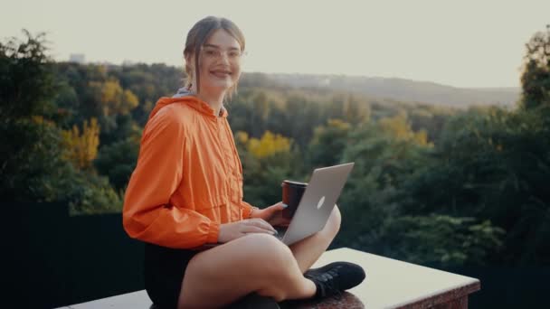 A vörös hajú, zsemlében ragadt fiatal nő a szoba elé néz, és széles mosollyal ül a parkban, a zöld fák hátterében. Egy laptopot tart a karjaiban és egy csésze kávét. — Stock videók