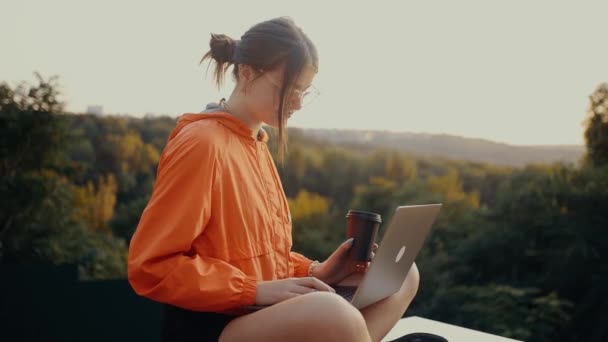 Het geconcentreerde meisje doet de boodschappen online, in het park, via de computer, met een kopje koffie in haar hand. Ze kijkt naar het laptopscherm, kijkt dan naar de camera en lacht breed. Koper — Stockvideo