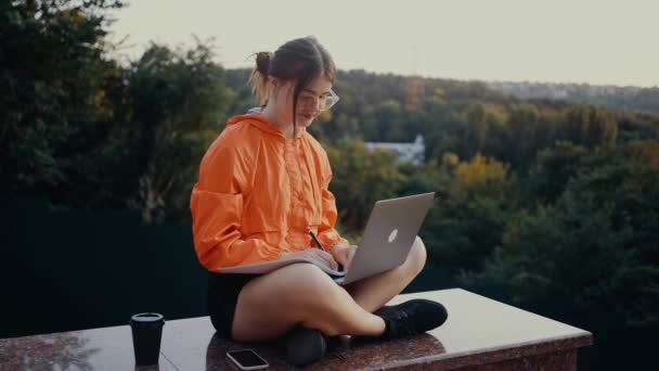 Die ehrgeizige Studentin macht sich Notizen aus dem Online-Kurs. Gekleidet in eine leuchtend orangefarbene Bluse, die Haare im Dutt, vor dem Hintergrund des grünen und dichten Waldes. 4k-Konzept — Stockvideo
