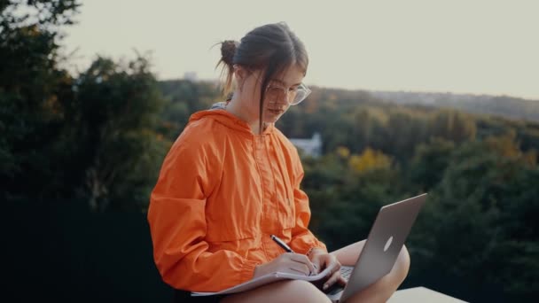 The young worker writes down her to-do list to achieve this months goals, in the middle of nature. Dressed in a bright orange blouse, hair caught in a bun, on the background of the green and dense — Stock Video