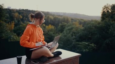 Çevrimiçi bir web sitesini izleyen ve doğanın ortasında not alan bir öğrencinin çerçevesi. Parlak turuncu bir bluz giymişti, saçları yeşil ve sık ormanın arka planında bir topuza sıkışmıştı. 4k
