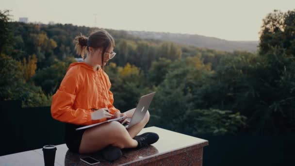 Keret egy diák, aki néz egy online webinar és jegyzetel, a természet közepén. Fényes narancssárga blúzba öltözve, zsemlébe akadt hajjal, a zöld és sűrű erdő hátterében. 4k — Stock videók