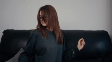 Genç kızıl saçlı, rahat, çilli ve gözlüklü, evdeki siyah kanepede oturarak dans hareketleri sergiliyor. Koyu yeşil bir bluz giymişti. Evde dinlenen kadın konsepti. 4k konsepti