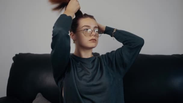 La joven pelirroja, pecosa, seria, con gafas coge su pelo en la cola. Vestido con una blusa verde oscuro, sentado en un sofá negro, en casa. El concepto de la mujer relajándose en casa. Concepto 4k — Vídeos de Stock