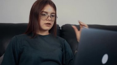 Gözlüklü kızıl saçlı kadın, online eğitim veren serbest çalışan. Koyu yeşil bir bluz giyip siyah bir kanepede oturuyor. Çalışan kadın konsepti. 4k konsepti