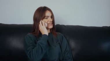 Kızıl saçlı mutlu bir kadın telefonda mutlu bir şekilde konuşuyor, önemli şeyleri tartışıyor. Yeşil bir bluz giymiş, evdeki siyah koltukta oturuyor. Sosyal adam kavramı. 4k konsepti