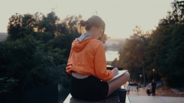 Genç kadın dizüstü bilgisayarıyla çalışırken ve not defterine not alırken, perspektifi kahve ile çalışan genç bir kadın tarafından filme alındı. Parlak turuncu bir bluz giymişti, saçları vardı.