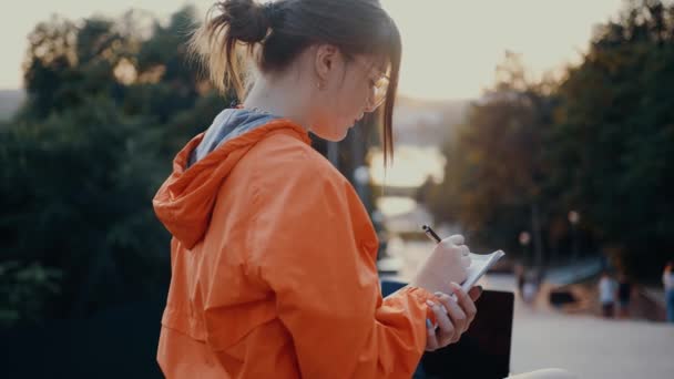 The young worker, filmed from behind, who takes notes and sets her list of goals for these months. Dressed in a bright orange blouse, hair caught in a bun, positioned on the park strip. 4k concept — Stock Video