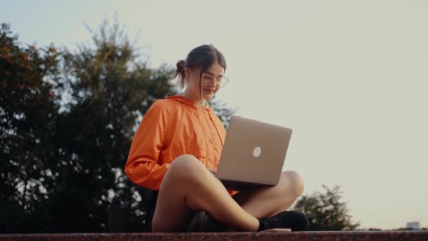 一位年轻的自由职业者正在她的笔记本电脑上做一个新项目 专心致志地打字 头抬起来 露出大大的笑容 穿着鲜艳的橙色衬衫 头发扎成一个发髻 穿着红色的衣服 — 图库视频影像