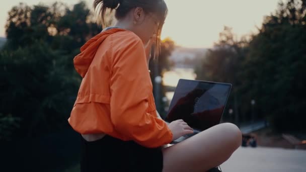 Egy gyönyörű, nyugodt fiatal nő ül a laptopján, és szeretne begépelni valamit. Fényes narancssárga blúzba öltözve, zsemlébe akadt hajjal, a park szélén. 4k koncepció — Stock videók
