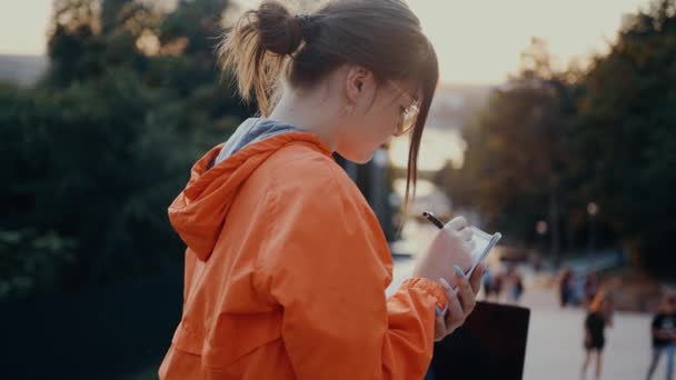 The ambitious student, filmed in profile, notes the subjects for the exam. Dressed in a bright orange blouse, hair caught in a bun, positioned on the park strip. 4k concept — Stock Video