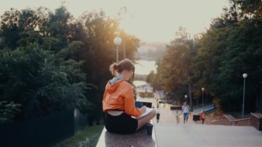 The ambitious young woman who takes notes from her laptop, takes a break, raises her hands and stretches. Dressed in a bright orange blouse, hair caught in a bun, positioned on the park strip. 4k