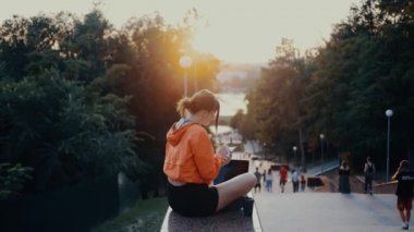 The young journalist writes a new article for the newspaper, makes notes, analyzes the world around her. Dressed in a bright orange blouse, hair caught in a bun, positioned on the park strip. 4k