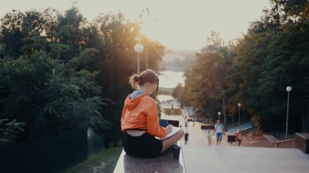 Амбітна молода жінка, яка робить нотатки зі свого ноутбука, робить перерву, піднімає руки і розтягується. Одягнена в яскраво-помаранчеву блузку, волосся спіймане в булочку, розташоване на парковій смузі. 4k — стокове відео