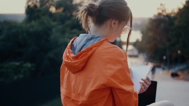 Young and hardworking woman makes notes, positioned on a bench in the park. Dressed in a bright orange blouse, hair caught in a bun, positioned on the park strip. 4k concept — Vídeo de Stock