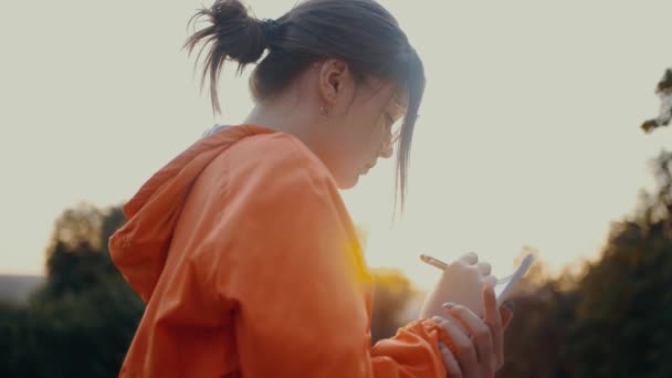 Close-up shot filmed from behind of an ambitious worker writing the plan for a new project. Dressed in a bright orange blouse, hair caught in a bun, positioned on the park strip. 4k concept — Αρχείο Βίντεο