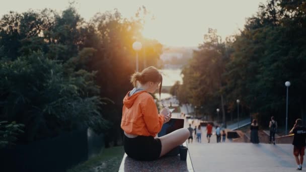 The young journalist writes a new article for the newspaper, makes notes, analyzes the world around her. Dressed in a bright orange blouse, hair caught in a bun, positioned on the park strip. 4k — 图库视频影像
