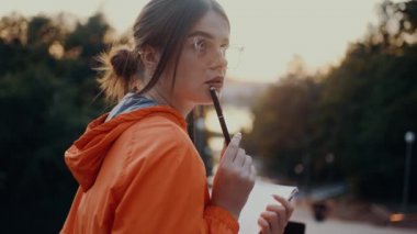A beautiful and creative young woman keeps her pen on her lips, she has a continuous idea to write in her notebook. Dressed in a bright orange blouse, hair caught in a bun, positioned on the park