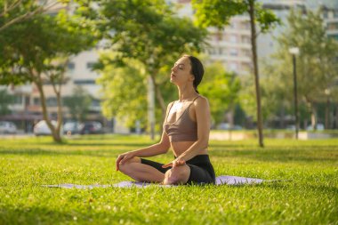 Mavi spor sutyenli ve siyah şortlu bir kadın yoga minderinde oturmuş göğsünde çimen zemininde ve arka planda bulanık bir elle maditasyon yapıyor..