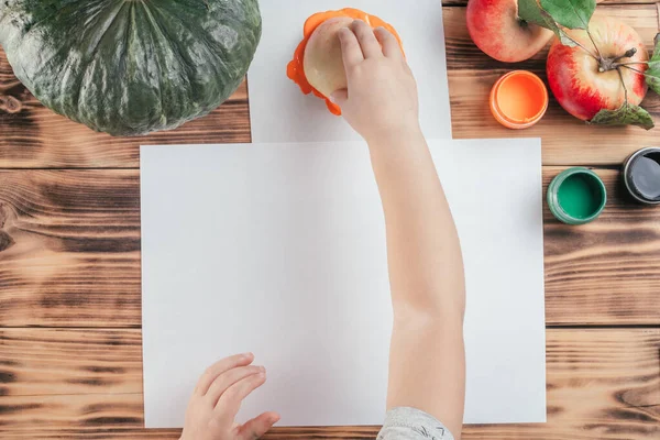 Adım adım Cadılar Bayramı çocukları özel balkabağı elması baskıları. Altıncı adım: Çocuk eli, elmanın yarısını turuncu guaj boyasına batırır. Üst görünüm