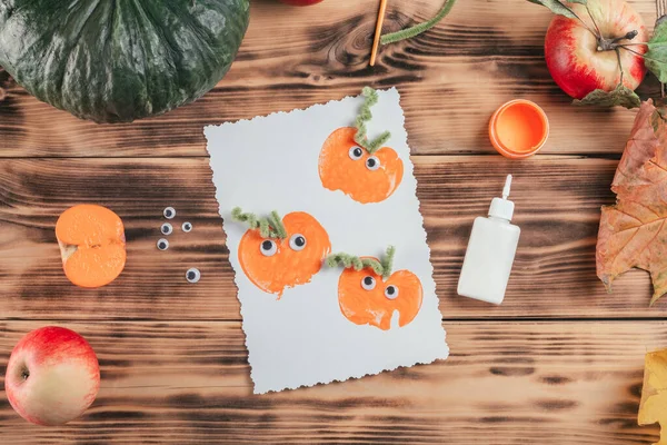 Finished Halloween card made with pumpkin apple prints lies next to glue and paint on wooden surface. Top view