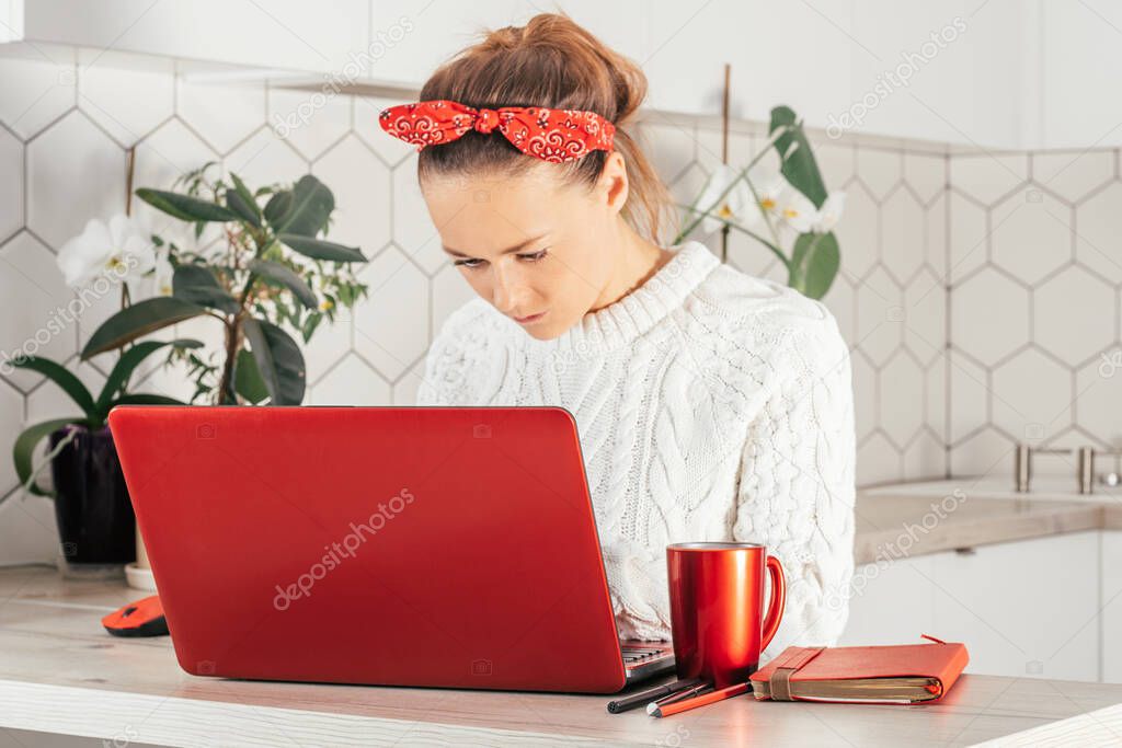 Young woman sits with red laptop at home in kitchen in red bandage and white sweater and looks at monitor. Home work or study concept