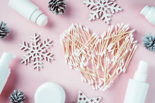 Wooden cotton swabs laid out in shape of heart with wooden snowflakes, pine cones, cosmetics jars and roll-on deodorant on pink background. Flat lay