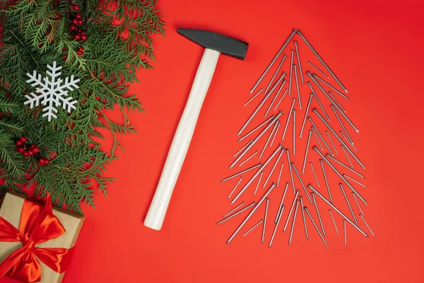Long nails and hammer, laid out in shape of Christmas tree with spruce branches and gift box on red background. Flat lay