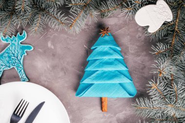 Noel ağacı ve tabağı şeklinde katlanmış keten peçeteler, bıçak, çatal ladin dallı gri beton yüzey, geyik ve eldiven seramik dekorasyonlar. Yeni yıl sofrası. Üst görünüm.