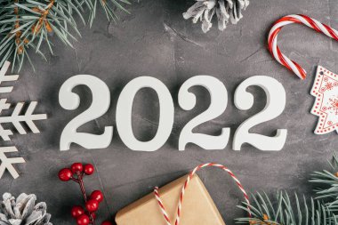 Mutlu yıllar 2022. Beyaz ahşap numaralar 2022. Noel ağacı dalları, süslemeleri, lolipopları ve kar taneleri ile gri düz olmayan beton yüzeyde yatıyorlar. Mutlu noeller. Üst görünüm