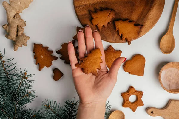 女性の手は クリスマスのモミの枝と白いテーブルの上の星やクリスマスツリーの形でクリスマスのジンジャーブレッドクッキーの上にクッキーを保持しています トップ表示 — ストック写真