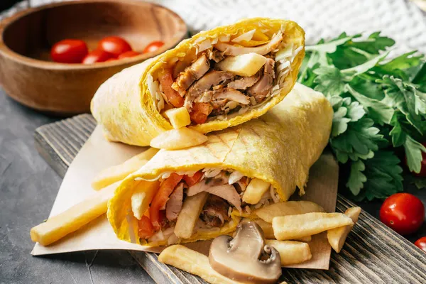 Rustic roll of shawarma with french fries, mushrooms, chicken and vegetables in pita bread on wooden dark background with cherry tomatoes and green parsley