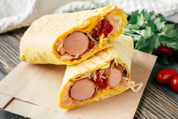 Roll with sausages and vegetables. Hot dog in pita bread on craft paper on wooden dark background with cherry tomatoes and green parsley