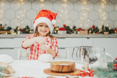 Kırmızı Noel şapkalı ve kareli gömlekli küçük siyah saçlı kız beyaz Noel süslemeli mutfakta çelenkli kurabiyeler için hamur hazırlıyor. Mutlu Noeller, yeni yıl.